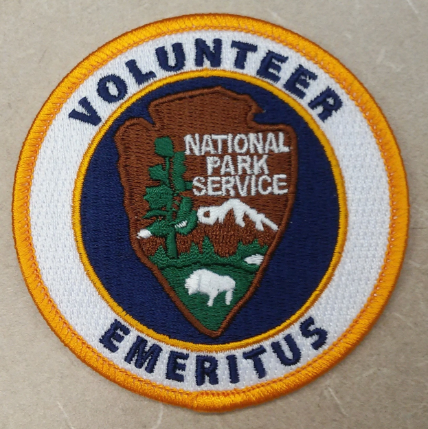 Runder Aufnäher mit blauem Rand und weißem Hintergrund, der in fetter schwarzer Schrift den Text "Volunteer National Park Service Emeritus" trägt, der in einem gelben Kreis in der Mitte platziert ist.