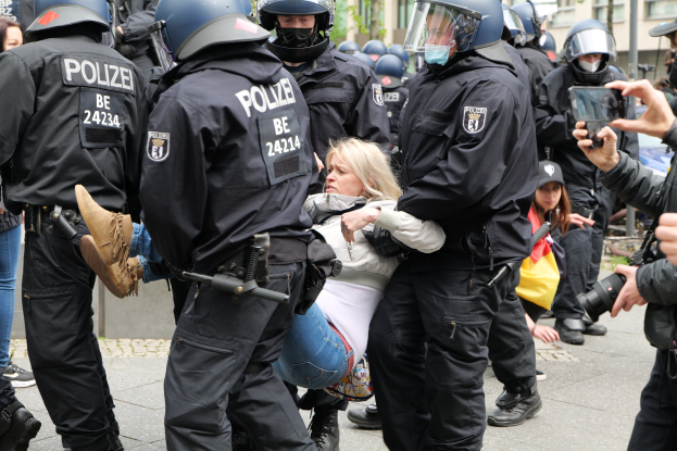 Polizeibeamte in Schutzausrüstung begleiten eine Frau in einem weißen und blauen Kleid, während ein Fotograf die Szene aufnimmt, mit Zuschauern und Gebäuden im Hintergrund.
