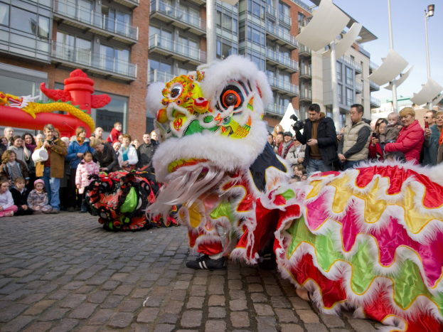 Ein lebendiges chinesisches Neujahrsfest in Amsterdam mit einem Drachentanz vor einem Publikum, das einige Kameras in der Hand hält, vor Gebäuden und Laternenmästen unter einem klaren blauen Himmel.