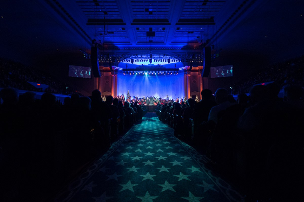 Ein großer Saal mit sitzenden Zuschauern, eine Bühne mit Musikern und Deckenleuchten und -fernseher, was auf einen Konzertsaal während der Nationalkonvention 2019 hinweist.