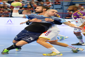 Eine Gruppe von Männern spielt Handball in einem Stadion, wobei einer den Ball hält und Zuschauer im Hintergrund sitzen.