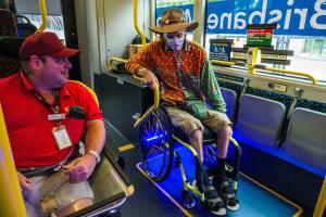 Zwei Männer im Rollstuhl in einem Bus, einer trägt ein rotes T-Shirt und eine rote Mütze, der andere ein orangefarbenes Hemd und einen braunen Hut, beide tragen Masken und Ausweise, mit gelben Stangen und Glasfenstern, die Bäume, Gebäude und Himmel zeigen.