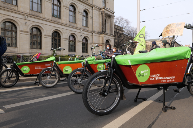 Eine Gruppe von Fahrrädern, die an einer Straße geparkt sind, mit einer Person in der Nähe, vor einem Gebäude, Bäumen und einem klaren Himmel, mit einem Fahrrad-Sharing-Banner im Vordergrund.