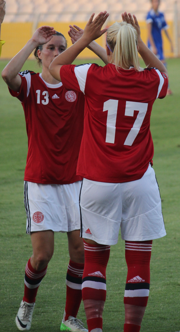 Zwei Frauen in roten T-Shirts und weißen Shorts stehen auf einem Fußballfeld und klatschen begeistert in die Hände, im Hintergrund eine Person in einem blauen Kleid, eine gelbe Tafel und ein paar Stühle.