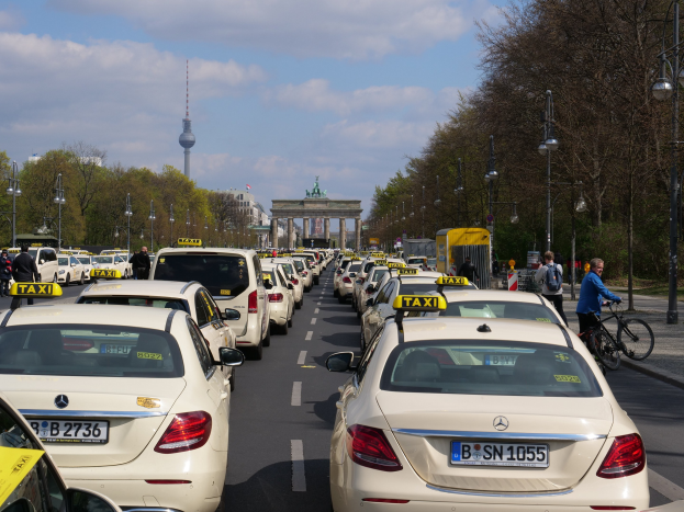 Eine lange Reihe von Taxis parkt entlang einer belebten Straße in Berlin, Deutschland, mit Fahrzeugen, Radfahrern, Fußgängern, Bäumen, Laternenmasten und Gebäuden im Hintergrund bei einem bewölkten Himmel.