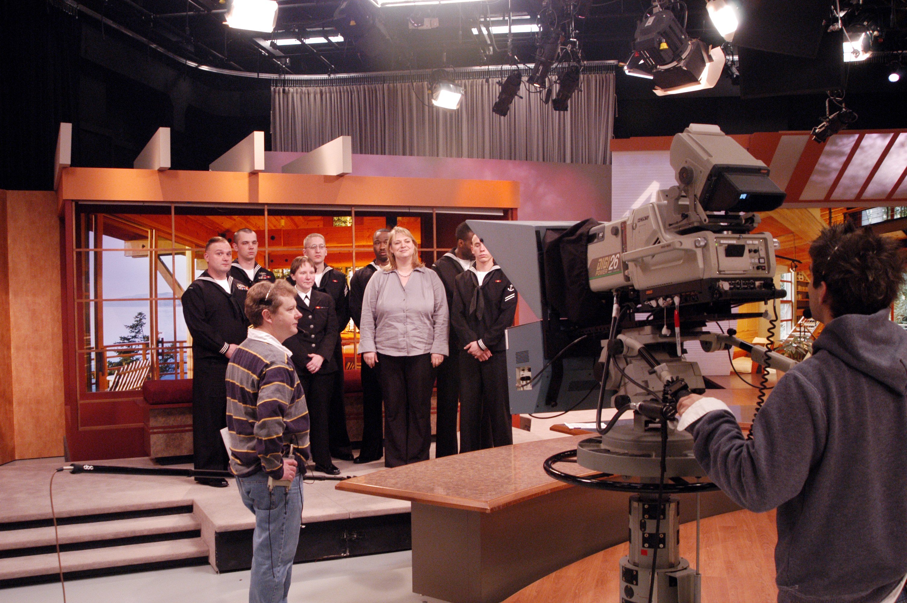 Gruppe von Menschen in einem Fernsehstudio mit einer Person, die eine Kamera hält, und Lampen an der Decke.