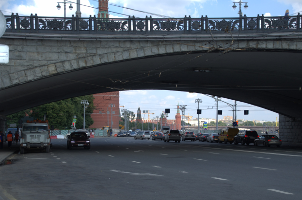 Eine Brücke überspannt eine vielbefahrene Straße mit darunter fahrenden Fahrzeugen, während Fußgänger auf dem Gehweg links stehen, mit städtischen Elementen wie Verkehrsampeln, Laternen, Bäumen, Gebäuden und Himmel im Hintergrund.