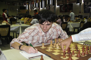 Mann bewegt Schachfigur und schreibt mit einem Stift, während im Hintergrund Menschen spielen.