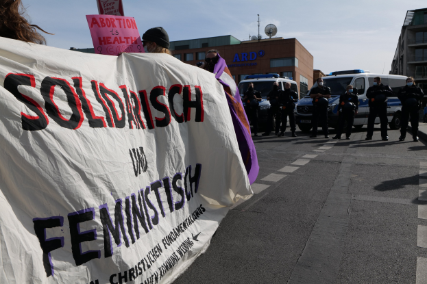 Eine Gruppe von Menschen marschiert auf einer Straße, hält eine "Solidarität und Feminismus"-Fahne, mit geparkten Fahrzeugen, Gebäuden, einer Schüsselantenne und einem klaren blauen Himmel im Hintergrund.