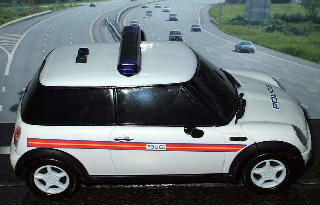 Weißer Polizeiwagen mit "Polizei" Aufschrift auf einer Straße vor einer Stadt, umgeben von Verkehr, Gras und Bäumen auf beiden Seiten.
