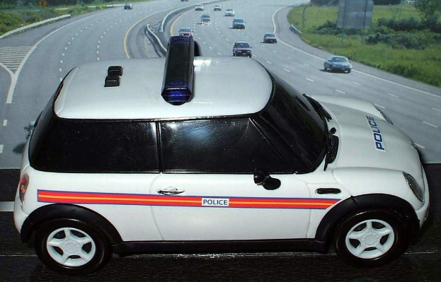 Weißer Polizeiwagen mit "Polizei" Aufschrift auf einer Straße vor einer Stadt, umgeben von Verkehr, Gras und Bäumen auf beiden Seiten.