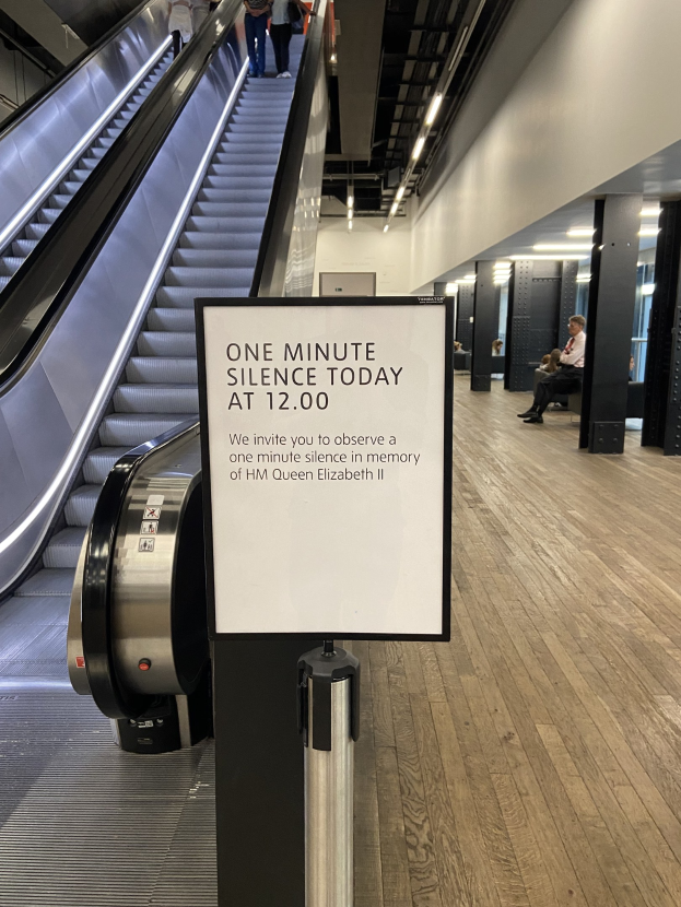 Eine Rolltreppe im Flughafen mit einem Schild, auf dem "Eine Minute Schweigen heute" steht, einige Menschen darauf und beleuchtete Decke im Hintergrund.