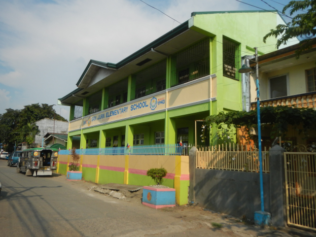 Ein Schulgebäude mit grüner und gelber Fassade, umgeben von einer Straße mit Fahrzeugen, Topfpflanzen, einem Zaun, einem Tor, einem Straßenschild, Bäumen, Drähten und einem bewölkten Himmel.