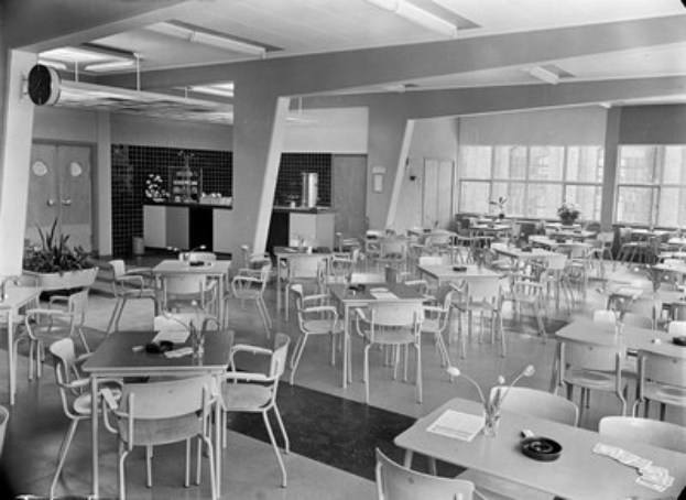 Schwarz-weißes Foto einer Cafeteria mit Tischen und Stühlen, Tellern, Besteck, Gläsern und Vasen auf den Tischen sowie Türen, Fenstern und Deckenleuchten im Hintergrund.