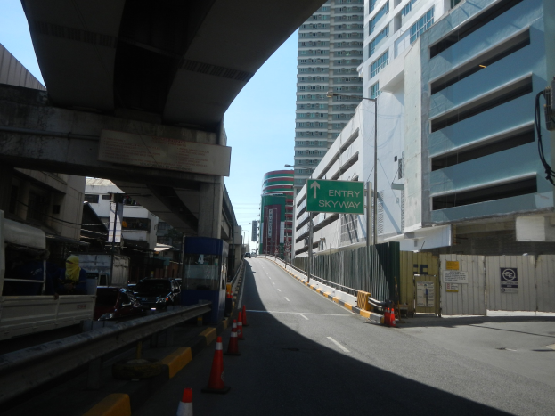 Stadtstraße mit Verkehrskegeln, Fahrzeugen, einer Brücke auf der linken Seite, Gebäuden mit Namensschildern, Laternenpfählen und einem Himmel im Hintergrund.