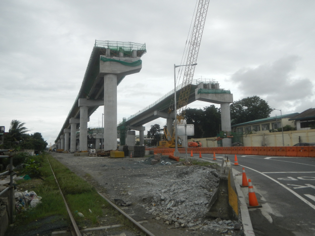 Baustelle mit einer Brücke im Hintergrund, Straße mit Absperrgittern markiert, Bahnschiene auf der linken Seite, verstreute Steine und Gras, Bäume und Gebäude säumen die Straße und ein bewölkter Himmel.
