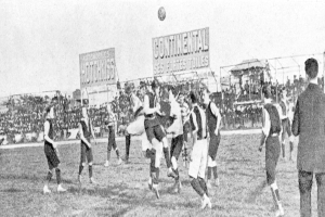 Ein Schwarz-Weiß-Foto einer Gruppe von Männern, die auf einem Feld Fußball spielen, mit einem Ball in der Luft und Zuschauern im Hintergrund, mit Schildern, die Text über das Spiel anzeigen.