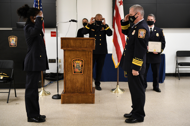 Eine Gruppe uniformierter und maskierter Personen steht vor einem Podium mit Mikrofon, umgeben von Fahnen, Stühlen und Tischen, während eine neue Feuerwache vereidigt wird, wobei eine Person ein Zertifikat hält.