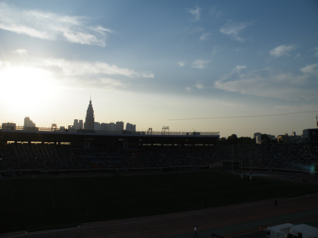 Ein Fußballfeld mit einem Stadion im Hintergrund, gefüllt mit Menschen, unter einem Himmel, der von Orangen- und Pinktönen zu Purpur übergeht, mit fernen Gebäuden, die sich gegen den Himmel abzeichnen.