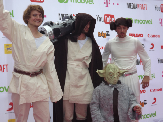 Eine Gruppe von Menschen in Star-Wars-Kostümen, die auf einem roten Teppich posen, wobei eine Person ein Objekt und ein Banner mit Text und Logos im Hintergrund hält.