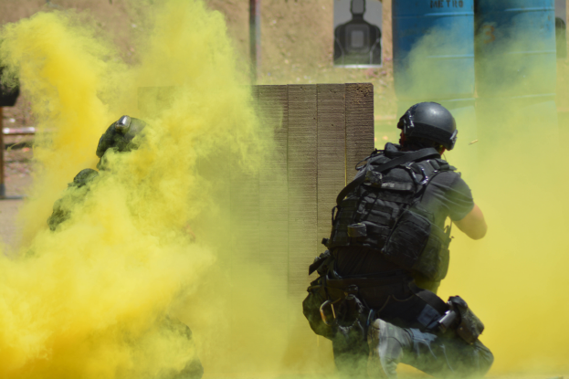 Zwei Polizisten in Einsatzausrüstung mit gelbem Rauch aus dem Mund, mit Helmen und Schutzausrüstung, vor einer Wand mit einer Tafel und anderen Gegenständen, mit dem Boden unten sichtbar.