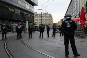 Gruppe von Polizisten in schwarzen Uniformen und Helmen auf einer Stadtstraße mit Glasfenstergebäuden, Bäumen und verstreuten Straßenmöbeln unter einem klaren Himmel.