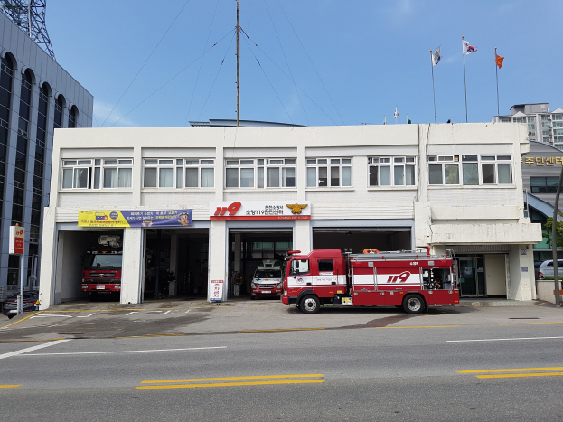 Feuerwache mit mehreren Feuerwehrautos vor dem Gebäude geparkt, ein Gebäude mit Fenstern und Bannern, Flaggen mit Stangen im Hintergrund, bewölkter Himmel und ein Turm auf der linken Seite.