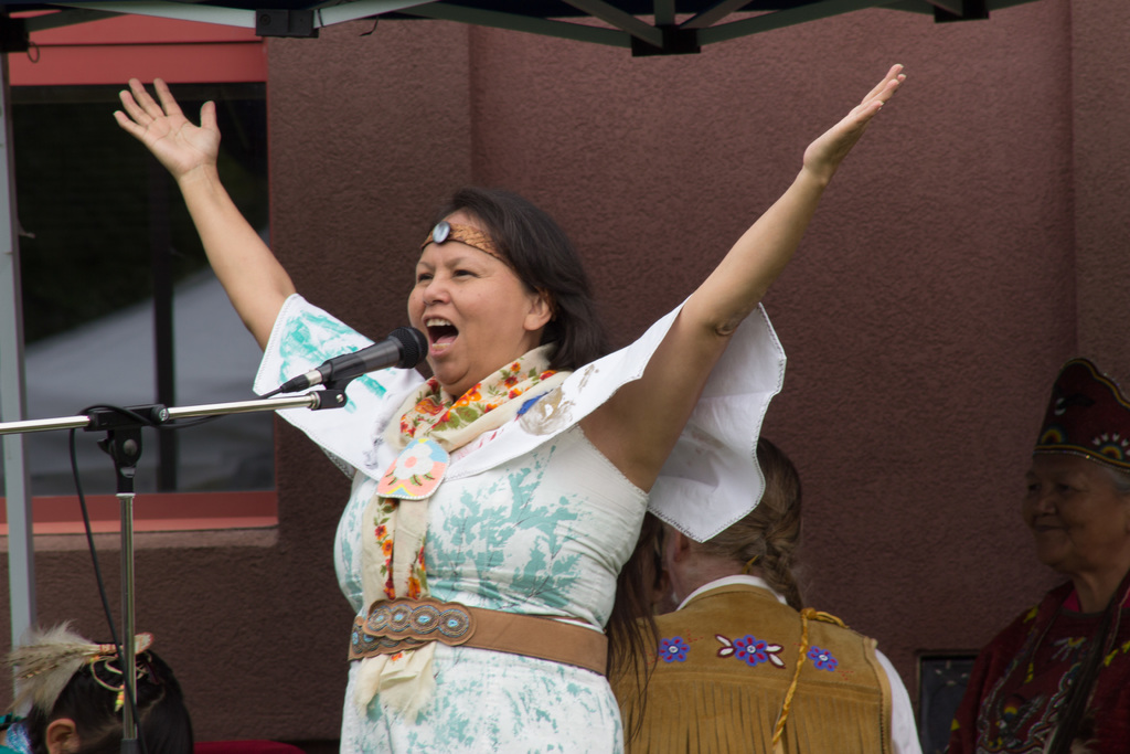 Eine Frau in einem weißen Kleid steht auf einer Bühne und spricht in ein Mikrofon, mit einer Wand und einem Eisenrahmen im Hintergrund.
