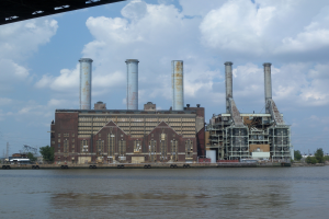 Ein großes Fabrikgebäude neben einem Gewässer mit einer Brücke und einem bewölkten Himmel im Hintergrund.