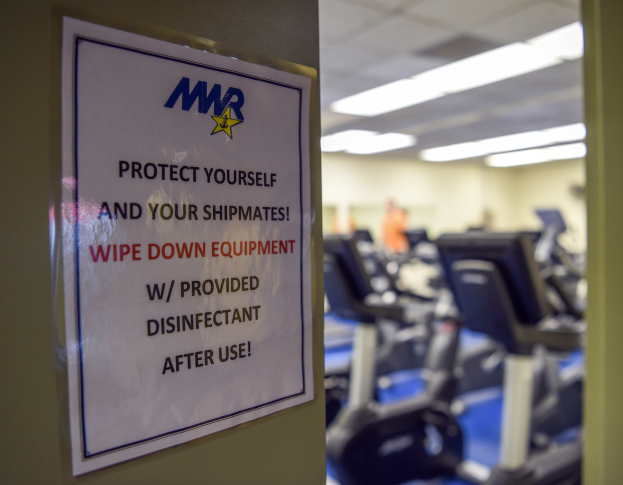 Zeichen an einer Turnhallentür mit der Aufschrift "Schützen Sie sich und Ihre Schiffskameraden" mit Laufbändern und Deckenleuchten im Hintergrund.