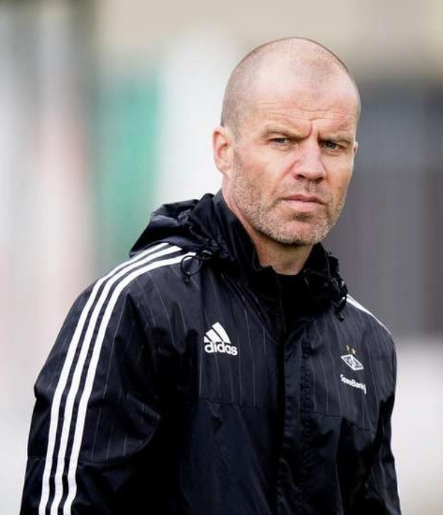 Ein kahlköpfiger Trainer in einer blauen Jacke mit weißen Streifen und einem weißen Kragen steht auf einem Fußballfeld mit verschränkten Händen und einem ernsten Gesichtsausdruck.