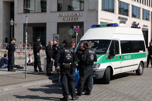 Eine Gruppe von Polizeibeamten steht vor einem Starbucks-Café mit einem Van auf der rechten Seite und Menschen auf der linken Seite, während im Hintergrund ein Gebäude mit Fenstern, ein Schild, ein Laternenpfahl und ein Zaun zu sehen ist.