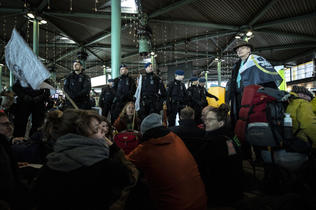 Eine Gruppe von Menschen steht vor einer größeren Menge in einem Bahnhof, mit Säulen und Deckenlampen im Hintergrund. Einige Personen in der Menge tragen Mützen, tragen Taschen und halten Schilder oder Banner, was auf eine Demonstration hindeutet.