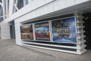 Großes Werbeplakat an einem Gebäude, das die Eröffnungszeremonie der Olympischen Spiele in Moskau ankündigt, mit sichtbarem Boden und rechter Beleuchtung.