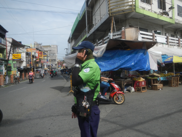 Ein Polizist in grüner und blauer Uniform und blauer Mütze steht an der Seite einer Straße und spricht auf einem Handy, mit Fahrzeugen im Hintergrund, Ständen mit verschiedenen Gegenständen rechts von ihm und einem bewölkten Himmel darüber.