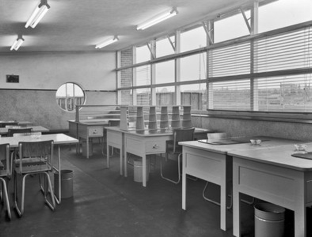 Schwarzes und weißes Foto eines Klassenzimmers mit Tischen, Stühlen, Bodenmülltonnen, einem wandmontierten Spiegel, Deckenleuchten und Fensterjalousien.