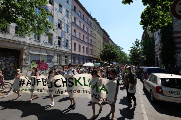 Eine Gruppe von Menschen marschiert auf einer Straße in Berlin, hält ein Banner in der Hand, mit einem Fahrradfahrer auf der linken Seite und parkenden Autos auf der rechten Seite, vor einem Hintergrund von Gebäuden, Bäumen und einem Schild.