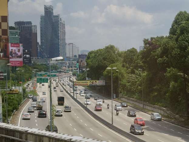 Eine belebte Autobahn mit hohen Gebäuden, Straßenlaternen, Schildern, Bäumen und einem bewölkten Himmel im Hintergrund.
