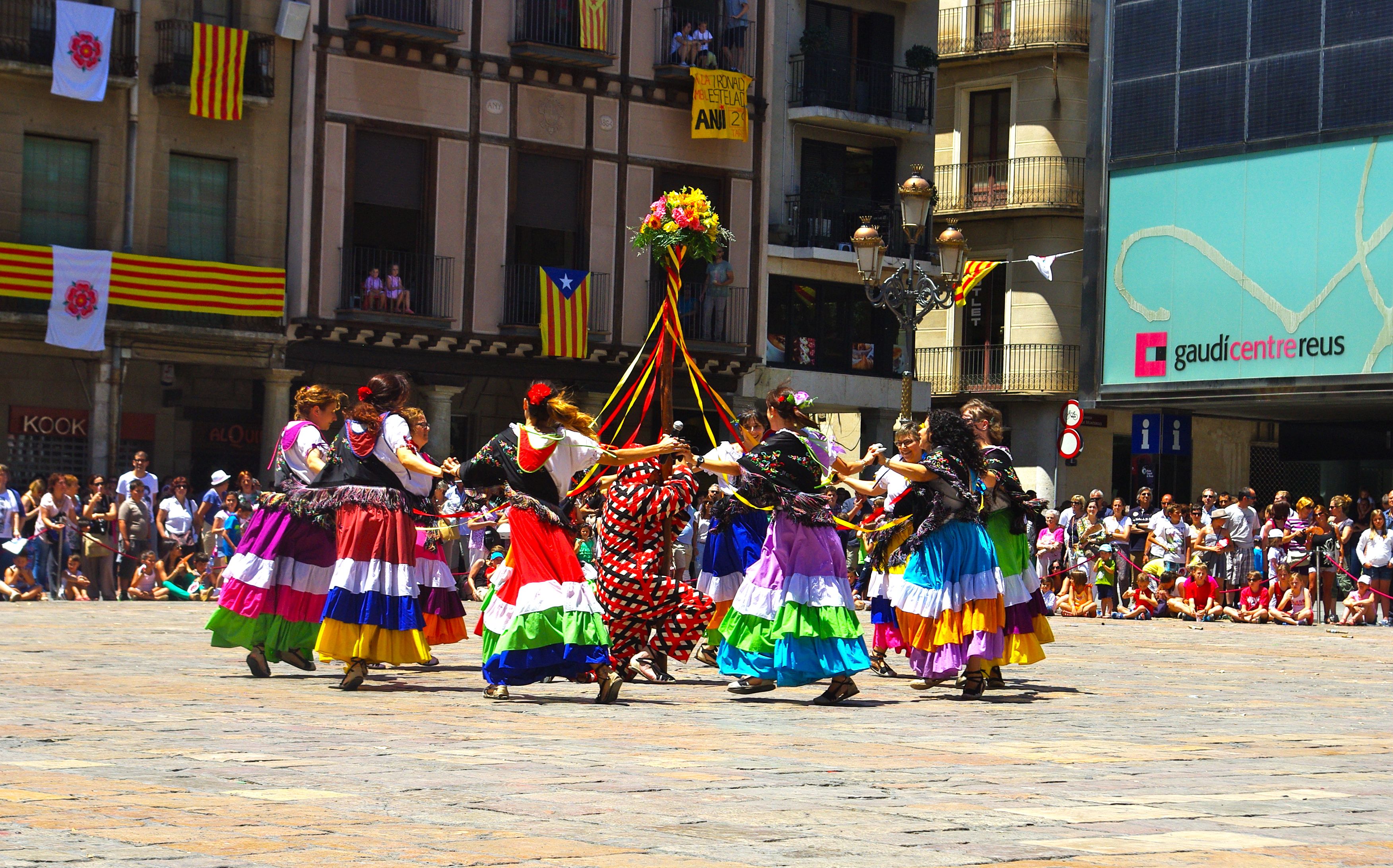 Eine Gruppe von Frauen in bunten Kleidern tanzt auf der Straße, umgeben von einer Menschenmenge, mit Gebäuden, Geländern, Balkonen, Fahnen, einem Laternenmast und einer Tafel mit Schrift im Hintergrund.
