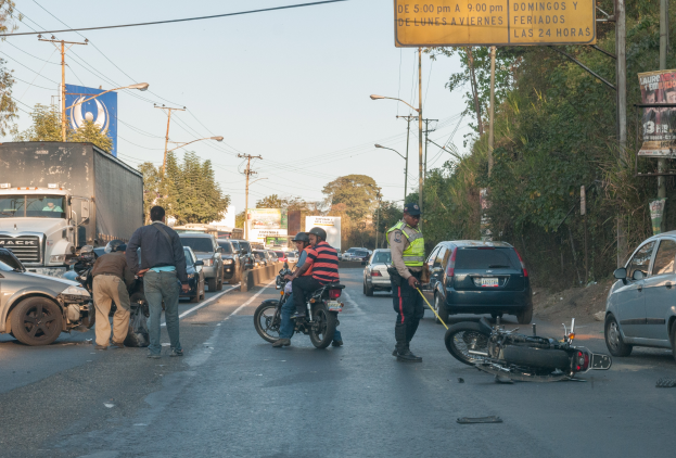 Eine Gruppe von Menschen um ein verunglücktes Motorrad auf der Straße herumstehend mit mehreren Fahrzeugen, darunter ein Lastwagen, und einem Hintergrund aus Bäumen, Pfählen, Lampen und Schildern unter dem Himmel.