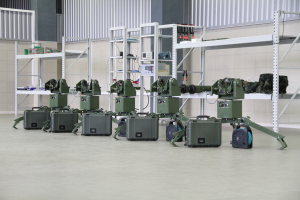 Eine Gruppe von Militärrobotern auf dem Boden, umgeben von Koffern und anderen Gegenständen auf Gestellen, mit einer Wand und einem Geländer im Hintergrund.