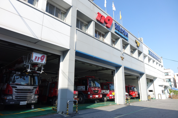 Feuerwache mit mehreren Feuerwehrautos vor einer Fensterfront mit Säulen, Namensschildern und Flaggen im Vordergrund vor einem Hintergrund aus Gebäuden und einem klaren blauen Himmel.
