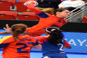 Eine Gruppe von Frauen beim Handballspielen in einer Arena, eine hält den Ball, Zuschauer sitzen im Hintergrund.