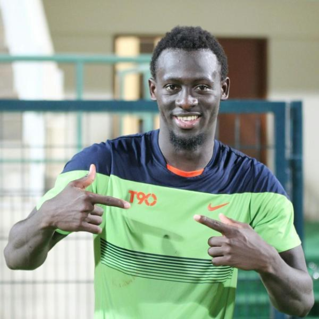 Ein Mann in einem grünen und blauen Hemd lächelt, während er auf die Kamera zeigt, mit einem unscharfen Geländer, einer Wand und einer Tür im Hintergrund.