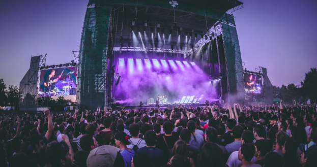 Eine große Menschenmenge versammelte sich auf einem Musikfestival, mit einer Bühne in der Mitte, auf der Musiker Instrumente spielten, beleuchtet von Lichtern und umgeben von Bildschirmen, mit einem Gebäude, Bäumen und einem klaren blauen Himmel im Hintergrund.