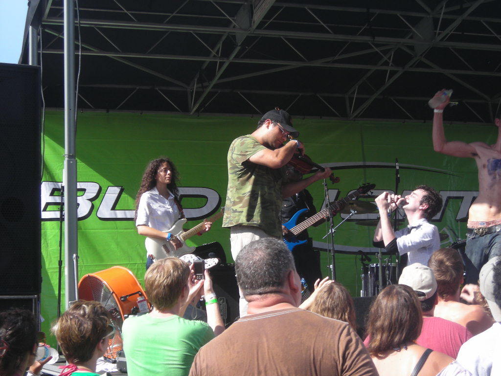 Eine Gruppe von Musikern performs auf der Bühne mit verschiedenen Instrumenten, während das Publikum vor einem Podium steht und die Musik genießt. Ein großer schwarzer Lautsprecher ist links positioniert und ein grünes Banner ist im Hintergrund sichtbar.