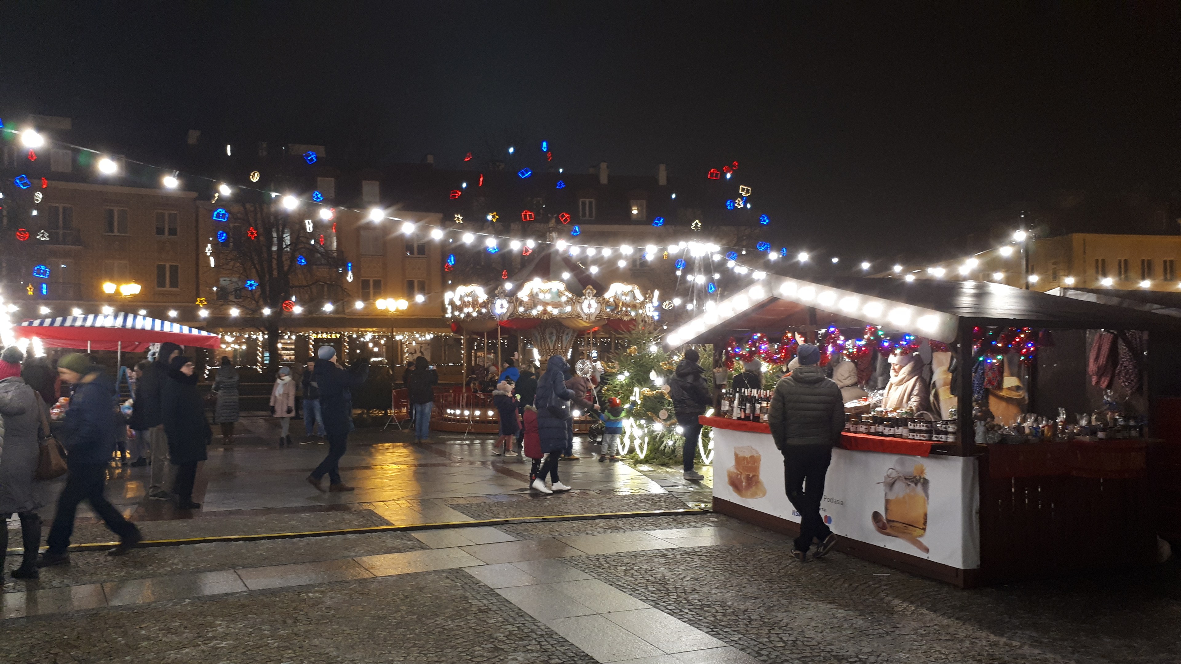 Ein belebter Weihnachtsmarkt bei Nacht in einer Stadt mit Menschen, beleuchteten Ständen, geschmückten Gebäuden, Bäumen und festlicher Beleuchtung am Himmel.
