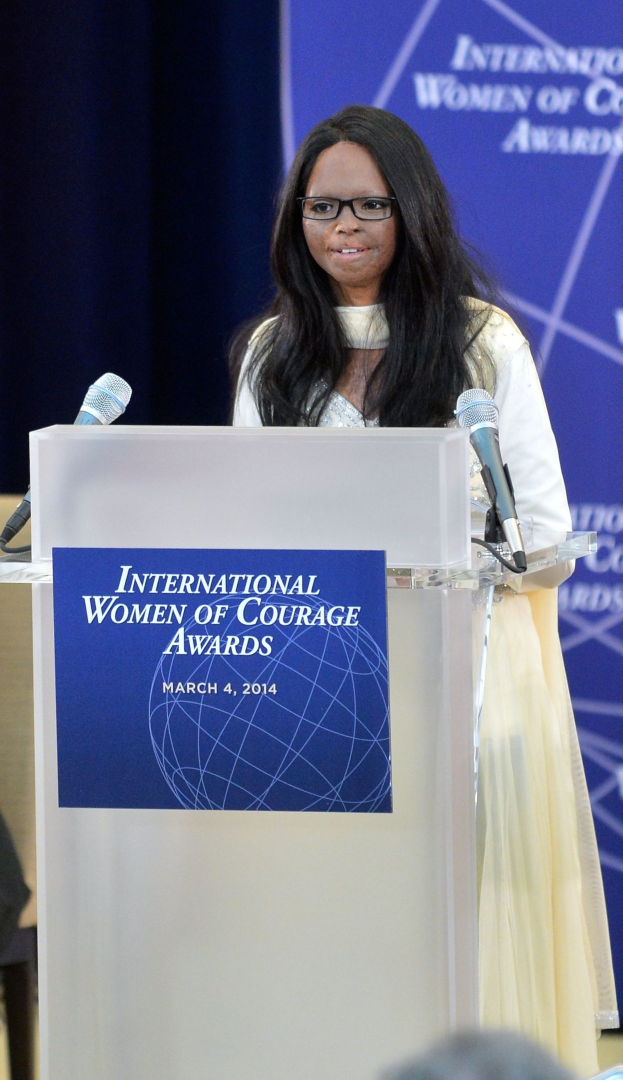 Frau in weißem Kleid und Brille, die an einem Pult mit zwei Mikrofonen und einer Tafel vor einem Publikum spricht, mit einem blauen Banner im Hintergrund, auf dem 'International Women of Courage Awards 2014' steht.