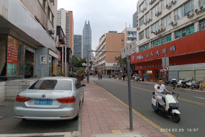 Ein Mann fährt auf einem Scooter eine Straße mit hohen Gebäuden entlang, mit Menschen auf den Bürgersteigen und Fahrzeugen auf der Straße, unter einem klaren blauen Himmel mit Bäumen im Hintergrund.