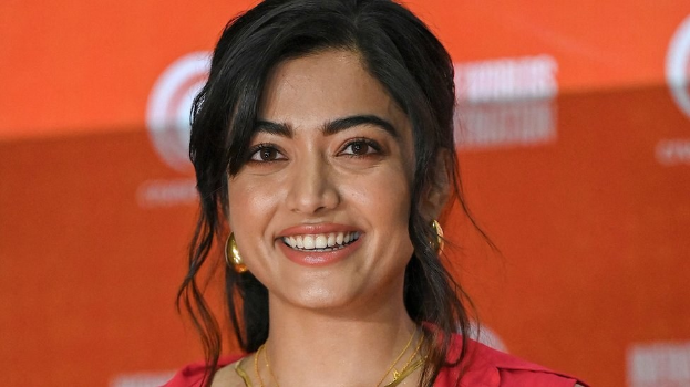 Eine Frau in einem roten Kleid mit einem eleganten Dutt und dezentem Make-up posiert strahlend vor der Kamera, trägt schwarze High Heels und hat einen hellgelben Banner mit der Aufschrift 'Sonam Kapoors Schönste Looks vom Cannes Film Festival' im Hintergrund.